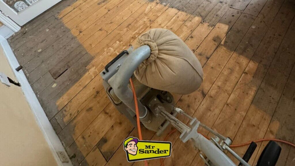 A floor sander removes layers of paint from a wooden floor, revealing the original wood grain. The sander has a dust collection bag attached to keep the area clean.