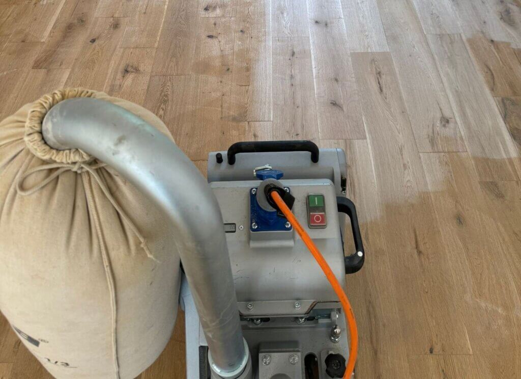 A professional from Mr Sander® is sanding a solid oak hardwood in a living room in Minley, UK using a Bona Belt sander (AMO110000.3). The machine features a 2.2 kW motor, 230 V voltage, 50 Hz frequency, and a sanding belt size of 200 x 750 mm or 250 x 750 mm. The total weight is 74 kg.