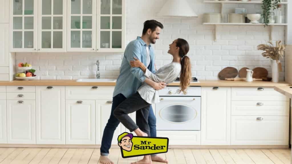 A joyful couple dancing in their beautifully renovated kitchen with hardwood floors by Mr Sander® in Clapham Junction, UK. The man dips the woman as they laugh, enjoying their time together. The kitchen features white cabinets, a white brick backsplash, and modern appliances, with the Mr Sander® logo visible at the bottom. The high-quality wood flooring adds warmth and elegance to the space, showcasing the exceptional floor sanding and refinishing services provided by Mr Sander® in Ringwould.