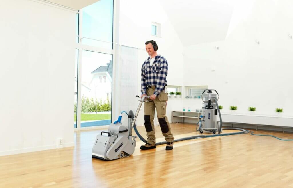A man from Mr Sander® is sanding a floor in a living room in the UK using professional machines, including a Bona Belt AMO110000.3 with a 2.2 kW motor, 230 V, 50 Hz, and a drum width of 200 & 250 mm. The room has green hanging plants, blue columns, and a bar with green tiles. Various tools and supplies are visible, indicating ongoing work. Dust containment is managed by a Bona DCS 70 with a 70 L capacity, HEPA fine filter, and 235 m³/hour airflow.