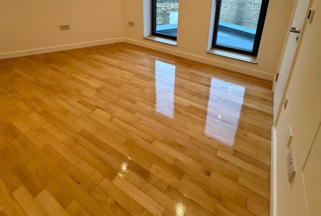 A freshly sanded and refinished light maple hardwood floor in a modern room. The floor has a high-gloss finish, reflecting light from the large windows with black frames. The natural grain of the maple wood is accentuated, creating a warm and polished look. The minimalistic room features clean white walls, allowing the beautiful wood floor to stand out.