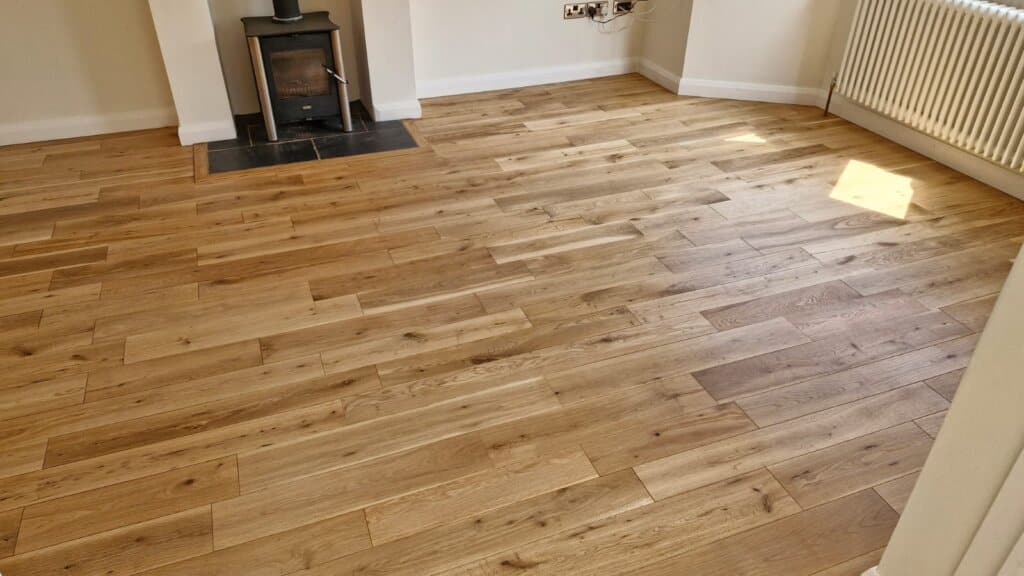 A beautifully refinished oak plank floor in a living room in Catford, UK. The floor, expertly sanded and refinished by Mr Sander®, showcases the natural grain and warm tones of the oak wood, enhancing the room's cozy and inviting atmosphere. The space features a modern wood-burning stove and light-colored walls, creating a comfortable and stylish setting.