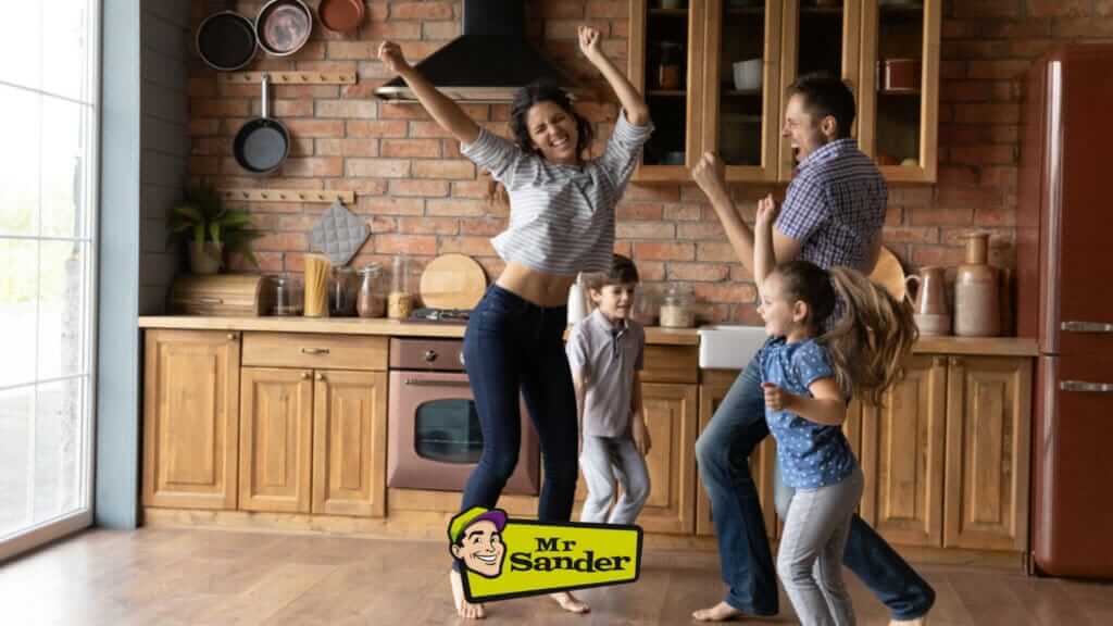 A joyful family dancing together in their beautifully restored kitchen with hardwood floors by Mr Sander® in Finchley Church End, UK. The parents and two children hold hands and spin around, smiling and laughing. The kitchen features rustic wooden cabinets, a brick backsplash, and modern appliances, with the Mr Sander® logo visible at the bottom. The expertly refinished floors add warmth and elegance to the home, showcasing the exceptional services provided by Mr Sander® in the Tonbridge area.