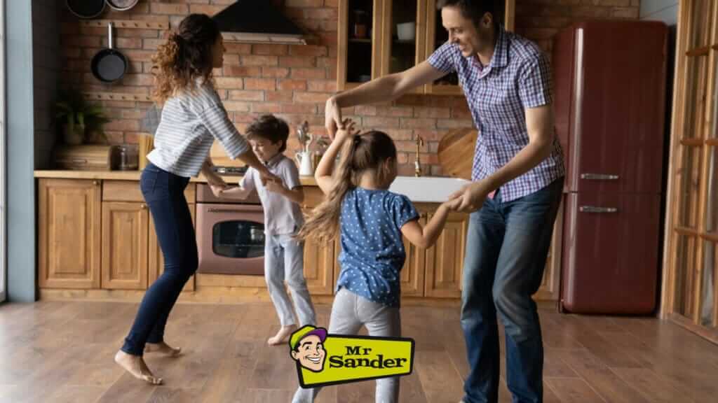 A joyful family dancing together in their beautifully restored kitchen with hardwood floors by Mr Sander® in Tonbridge, UK. The parents and two children hold hands and spin around, smiling and laughing. The kitchen features rustic wooden cabinets, a brick backsplash, and modern appliances, with the Mr Sander® logo visible at the bottom. The expertly refinished floors add warmth and elegance to the home, showcasing the exceptional services provided by Mr Sander® in the Tonbridge area.
