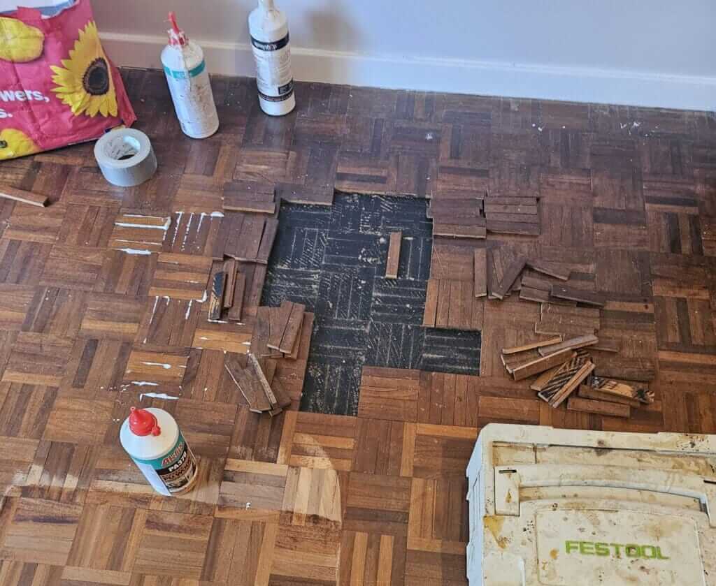 A parquet floor in the middle of a repair, with several tiles removed, revealing the black underlayment underneath. Glue and filler bottles, along with various tools and parquet pieces, are scattered around. Some of the parquet tiles are stacked nearby, ready to be installed to fill the missing gaps. A toolbox and a bag of materials are visible, indicating ongoing restoration work.