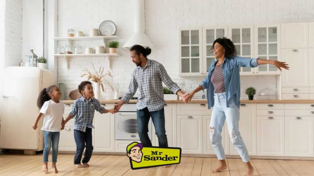 A happy family dances in their kitchen with a beautifully refinished pine plank floor in Aylesbury, UK. The floor, expertly sanded and refinished by Mr Sander®, complements the white cabinetry and modern decor, creating a bright and cheerful atmosphere. The family’s joy and satisfaction reflect the quality of the floor restoration.