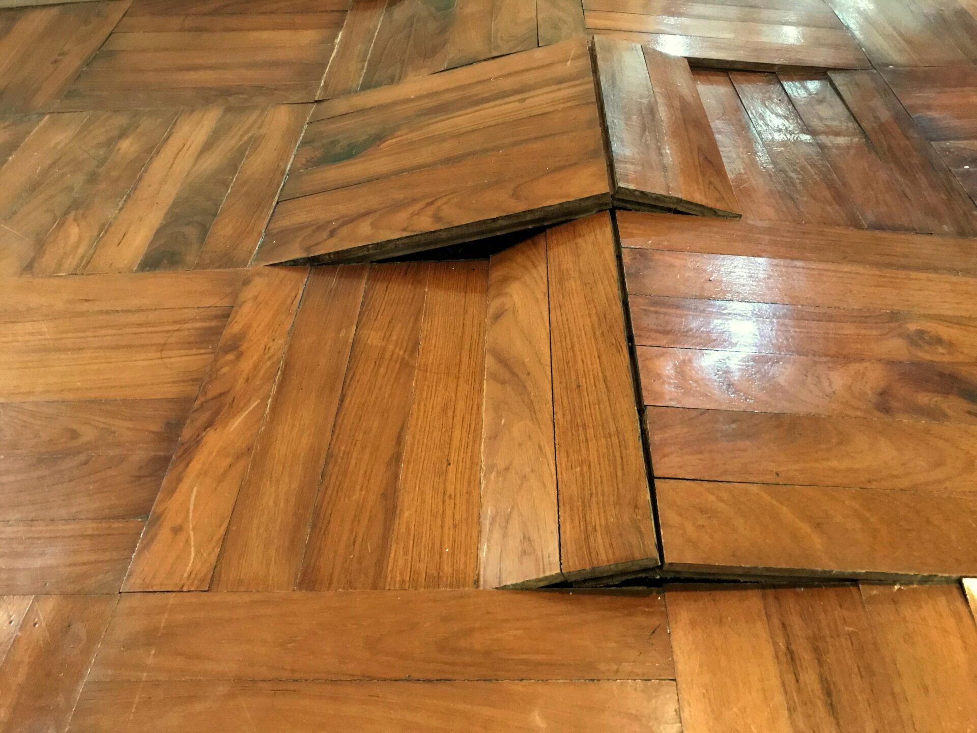 A close-up of a parquet wooden floor with a section raised and damaged, indicating a need for repair.