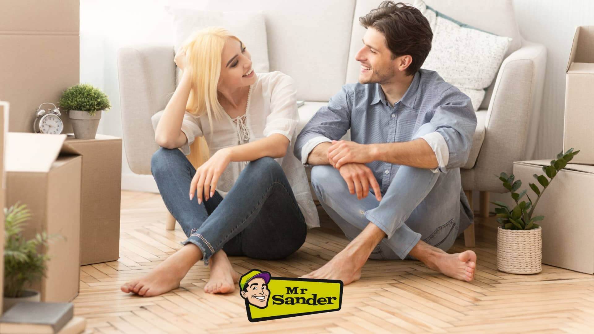 A happy couple sitting on the floor of a bright room with newly sanded herringbone wooden flooring, surrounded by moving boxes and plants. The "Mr. Sander" logo is at the bottom of the image.