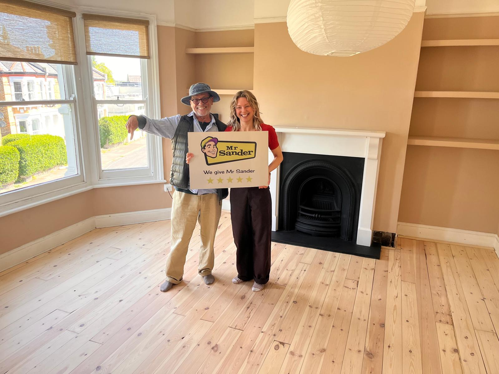 A satisfied customers holding a "Mr Sander" sign stands in a room with newly refinished wooden flooring, a black fireplace, white walls, and a window with white shutters.