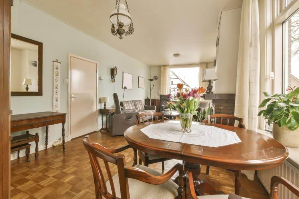 A cozy living and dining room featuring a beautifully restored parquet floor. The room is decorated with classic wooden furniture, including a round dining table with a floral centerpiece and cushioned chairs. Large windows let in natural light, complementing the warm tones of the parquet flooring. The space has a traditional and inviting atmosphere, with soft pastel walls and elegant décor.