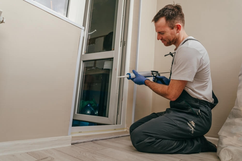 A man kneeling on the floor, using a caulking gun to fill gaps near a sliding door in a room. He is wearing gloves and work overalls, focusing on precise application of the sealant.