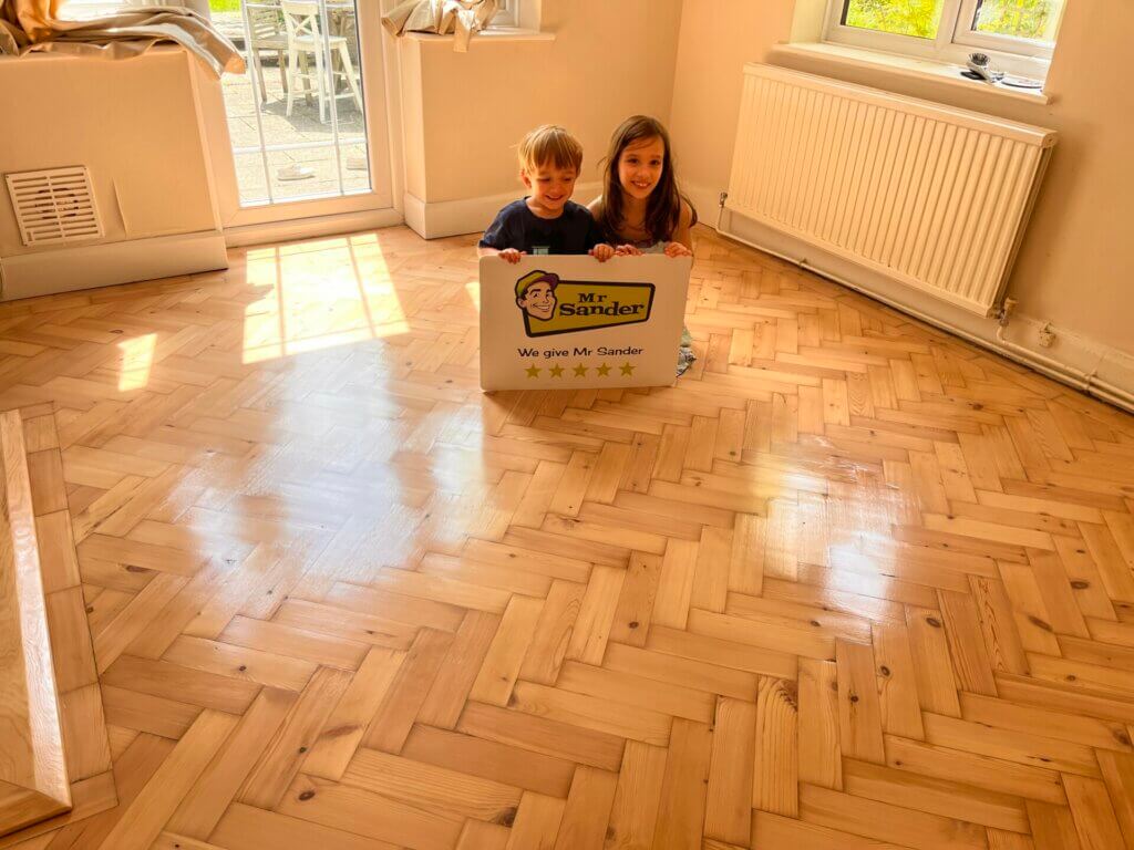 Two children sitting on a newly refinished floor in UK, holding a sign that reads "Mr Sander® - We give Mr Sander 5 stars". The room features beautifully sanded and refinished herringbone-patterned hardwood floors, a white radiator, and large windows letting in natural light.
