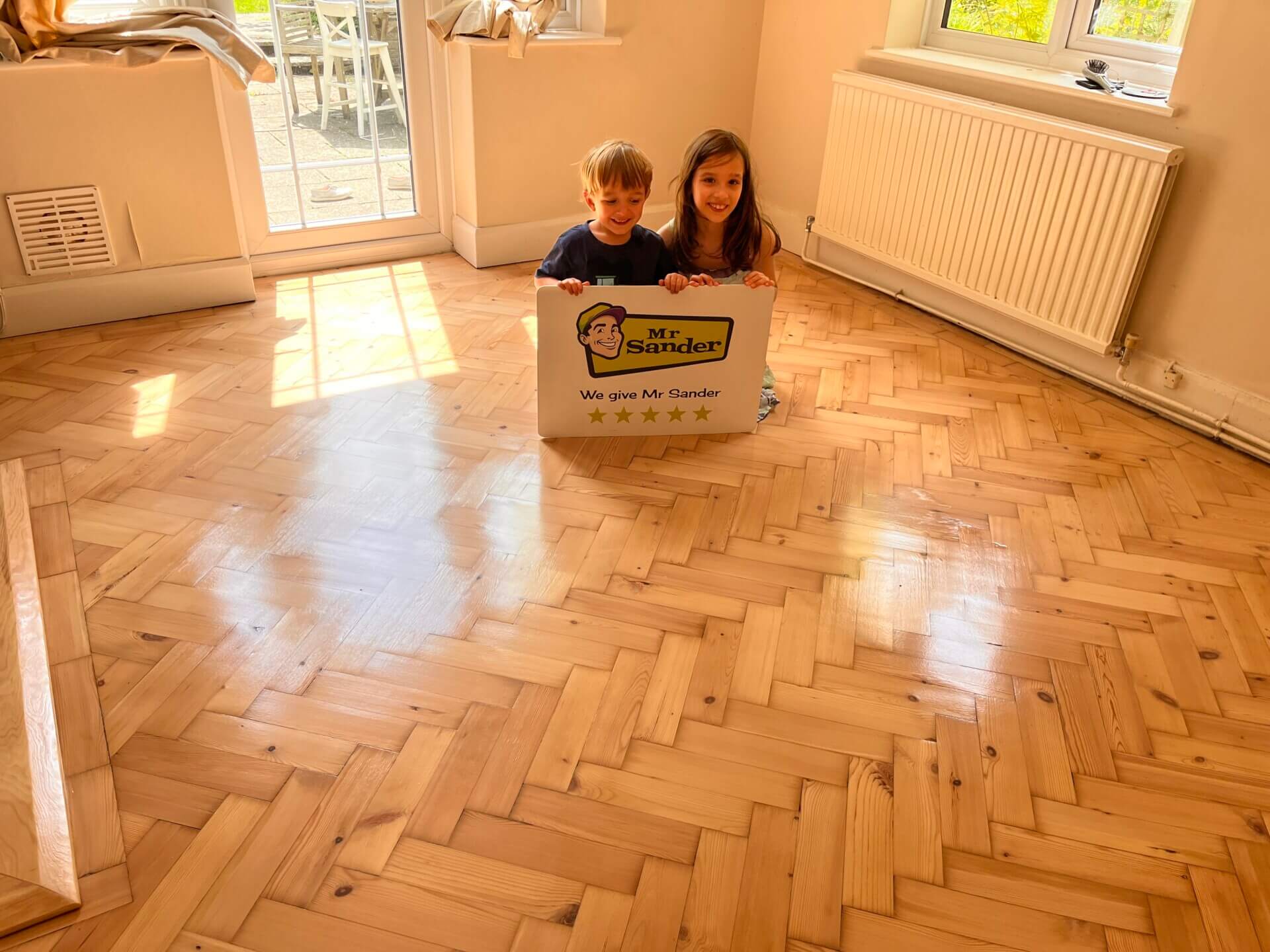 Two children sitting on a newly refinished floor in UK, holding a sign that reads "Mr Sander® - We give Mr Sander 5 stars". The room features beautifully sanded and refinished herringbone-patterned hardwood floors, a white radiator, and large windows letting in natural light.