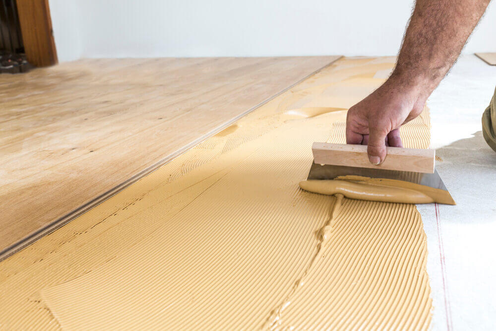 A flooring professional spreads adhesive on a prepared subfloor before installing wooden floor panels - East London Floor Fitting