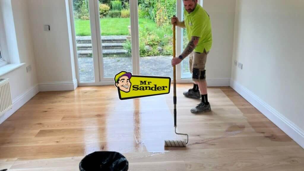A professional applying a finish to a wooden floor using a roller in a bright room with a garden view. The Mr Sander® logo is displayed prominently.