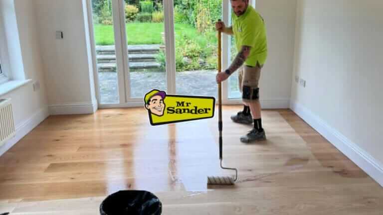 A professional applying a finish to a wooden floor using a roller in a bright room with a garden view. The Mr Sander® logo is displayed prominently.