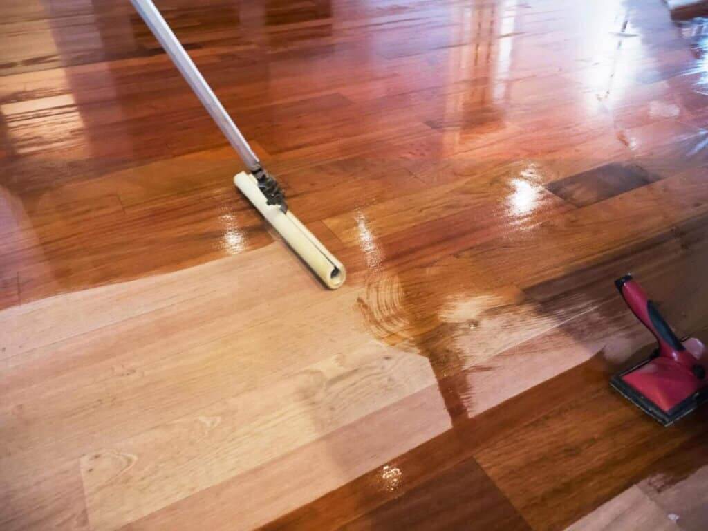 A close-up view of a hardwood floor during the finishing process, showcasing the application of a glossy oil finish. The image features a mop applicator smoothing the finish onto the floor, with one section already gleaming and another still in the application stage. The floor displays various shades of wood, indicating the richness of the natural grain, likely from species such as mahogany or cherry, highlighting the meticulous work of Mr. Sander® in the sanding and refinishing process.