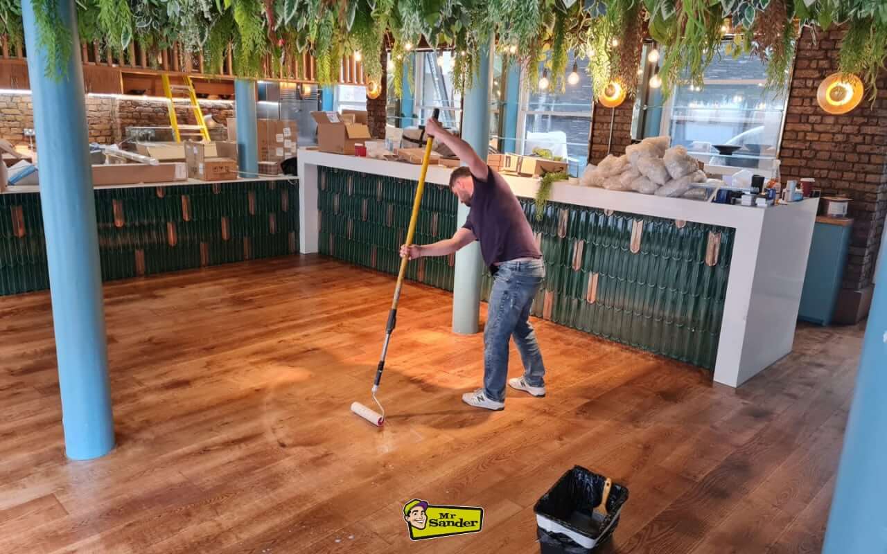 A professional floor refinisher stands in a newly renovated commercial space, rolling a protective finish onto the wooden floor.