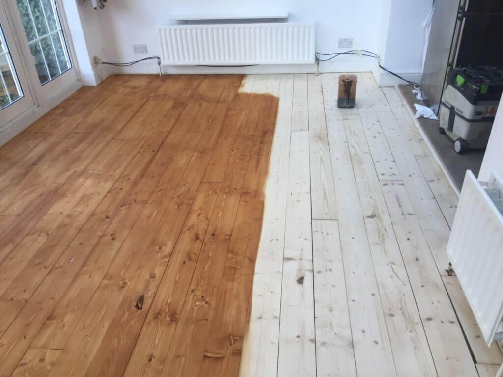 A room with wooden flooring undergoing staining. The left side of the floor is fully stained a warm, honey color, while the right side remains unstained, showing the natural light color of the wood. Various tools and equipment are scattered around, and there is a noticeable difference between the finished and unfinished areas.
