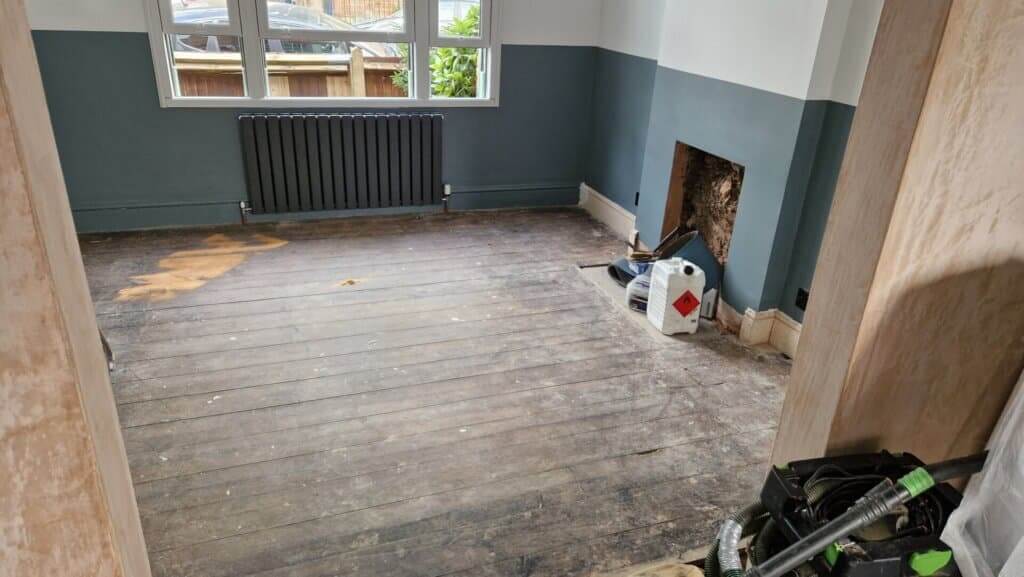 A room with old, worn wooden flooring that shows signs of heavy use and damage. The floor is dark and uneven, with visible scratches and stains. The room has dark grey walls, a black radiator under the window, and an unfinished fireplace area. Tools and materials are visible, indicating that restoration work is about to begin.