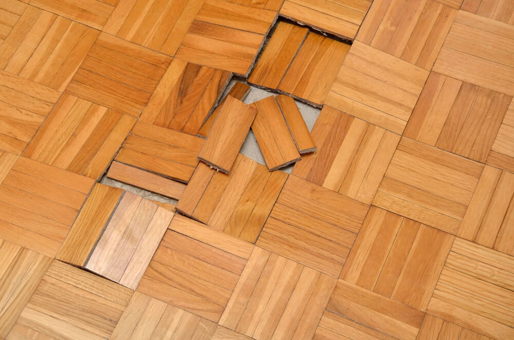 A close-up of a damaged wooden parquet floor, with several loose and displaced wooden tiles. The underlying subfloor is visible in the damaged area, indicating the need for repair.