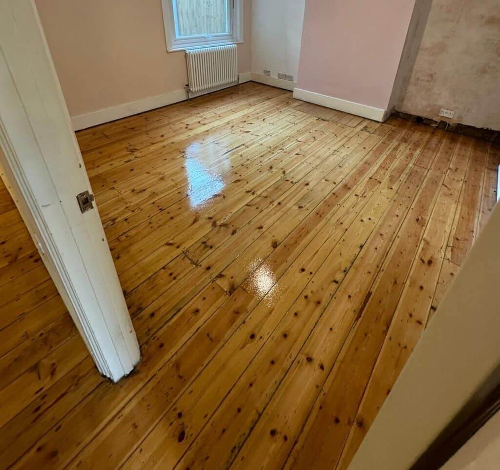 A room with freshly refinished wooden flooring, showcasing natural pine wood with visible knots and grain. The floor has a shiny, polished finish, reflecting light from a window. The room features pale pink walls, a white radiator beneath the window, and a partially visible open door.