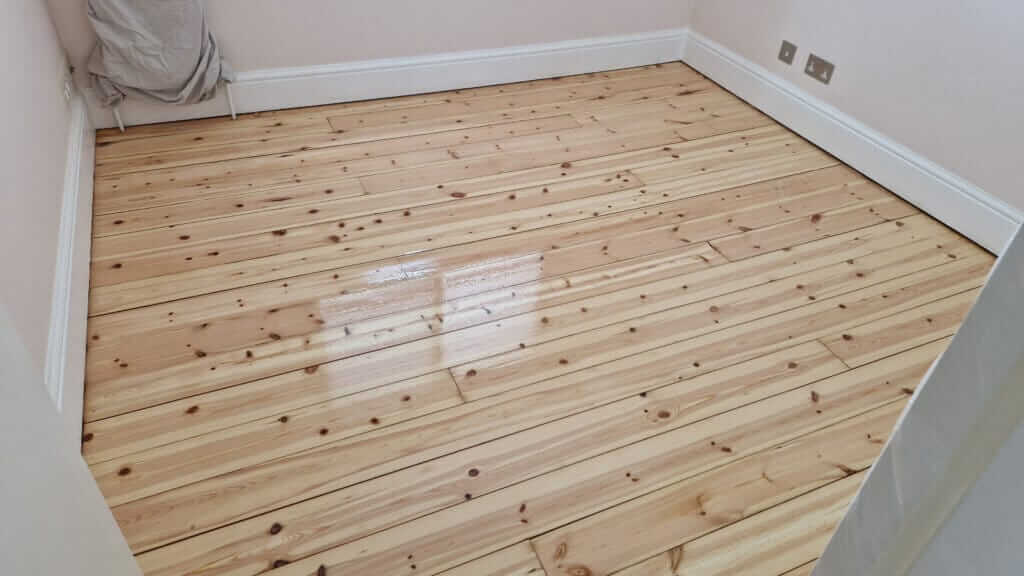 A beautifully restored pine wood floor in a room, showing a smooth, polished finish with sealed gaps between the boards. The floor reflects light, indicating a high-quality sanding and sealing job.