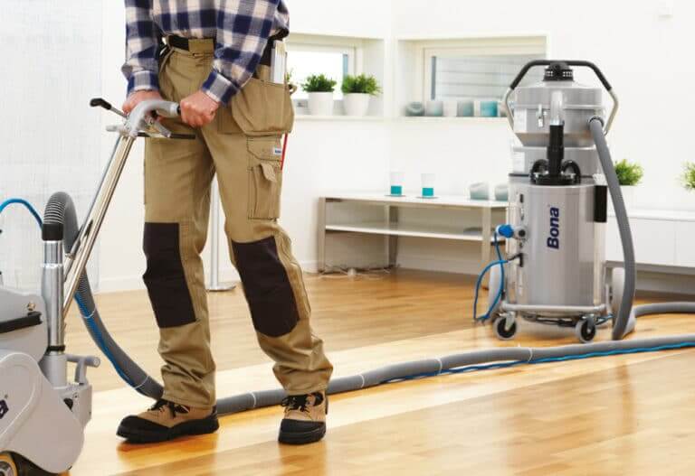 A technician from Mr. Sander® is sanding an oak wood floor in a living room, using a Bona Belt (AMO110000.3) machine. The professional belt sander features a motor power of 2.2 kW, operates at 230 V and 50 Hz, with a sanding belt size of 200 x 750 mm or 250 x 750 mm. The machine has a drum width of 200 & 250 mm and a total weight of 74 kg. Additionally, the Bona DCS 70 dust containment system is used, featuring a 70 L dust holding capacity, HEPA filter, and 7.5 m hose, ensuring a clean and efficient sanding process.