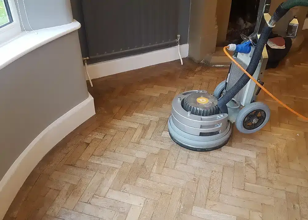 Image depicts a man from Mr Sander® sanding a parquet floor in a East Acton, UK living room. He operates the PALLMANN SPIDER Wood Floor Sanding Machine, featuring 360° illumination, switchable power reserve, and triple-disc technology. With 230V power supply, 2000W consumption, 50-400 RPM rotation speed, 42kg weight, 400mm sanding width, and 15kg additional weight.
