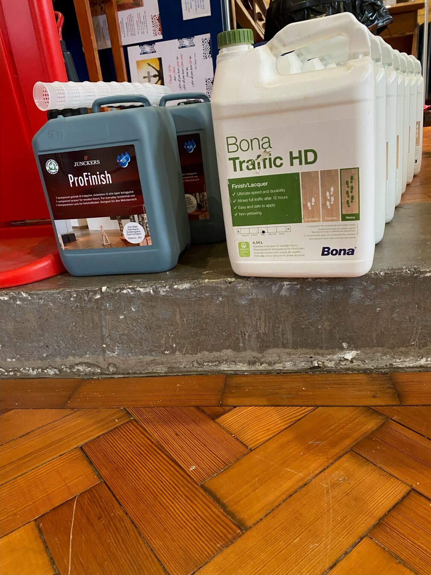 Bona Traffic HD and Junckers ProFinish lacquer containers standing on a step beside a partially sanded parquet floor.