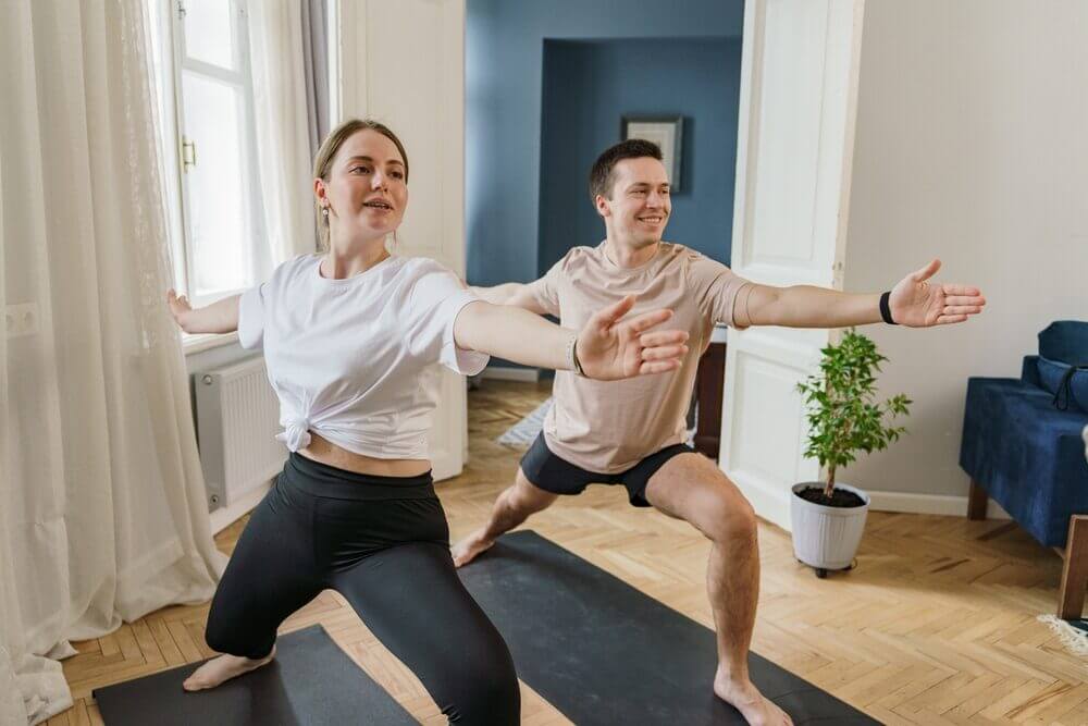A smiling couple performing yoga poses on a freshly installed herringbone wood floor, illustrating the comfort and style of a West London Floor Fitting.
