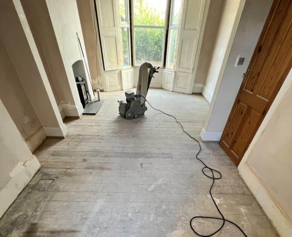 A professional from Mr Sander® is sanding a solid oak hardwood in a living room in UK using a Bona Belt sander (AMO110000.3). The machine features a 2.2 kW motor, 230 V voltage, 50 Hz frequency, and a sanding belt size of 200 x 750 mm or 250 x 750 mm. The total weight is 74 kg.