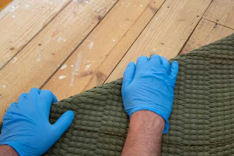 A pair of blue-gloved hands lifting a green carpet to reveal wooden floorboards beneath, highlighting a carpet to hardwood transformation.