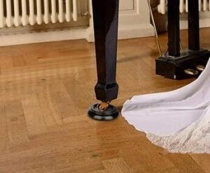 A black piano leg resting on a piano caster cup on a wooden floor, with a white cloth partially visible nearby.
