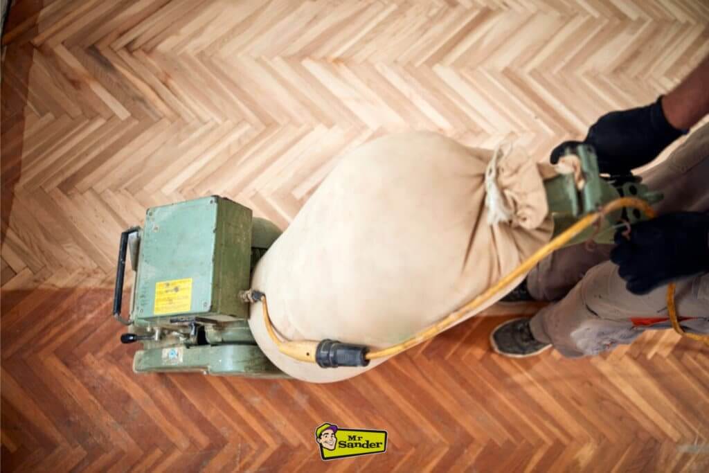 A professional from Mr Sander using a floor sanding machine to restore a herringbone parquet floor in progress. The top-down view captures the transformation between the sanded and unsanded sections.