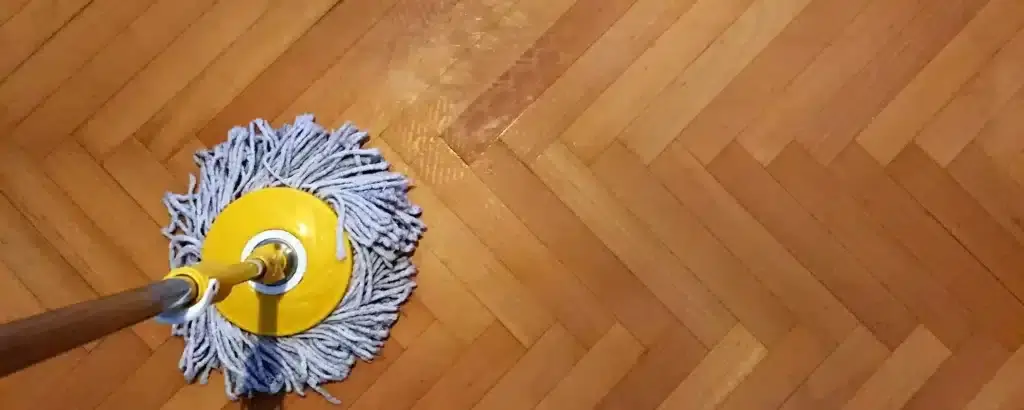 A close-up view of a herringbone parquet floor being cleaned with a yellow mop. The floor has a smooth, glossy finish, showcasing the quality of the sanding and refinishing work done by Mr Sander®.