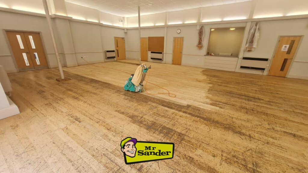 A large room with a wooden floor undergoing sanding and refinishing. A floor sanding machine is positioned in the center of the room, partially sanded and unsanded sections of the floor visible. The room features neutral walls, wooden doors, and several covered items. The Mr Sander® logo is placed at the bottom of the image.