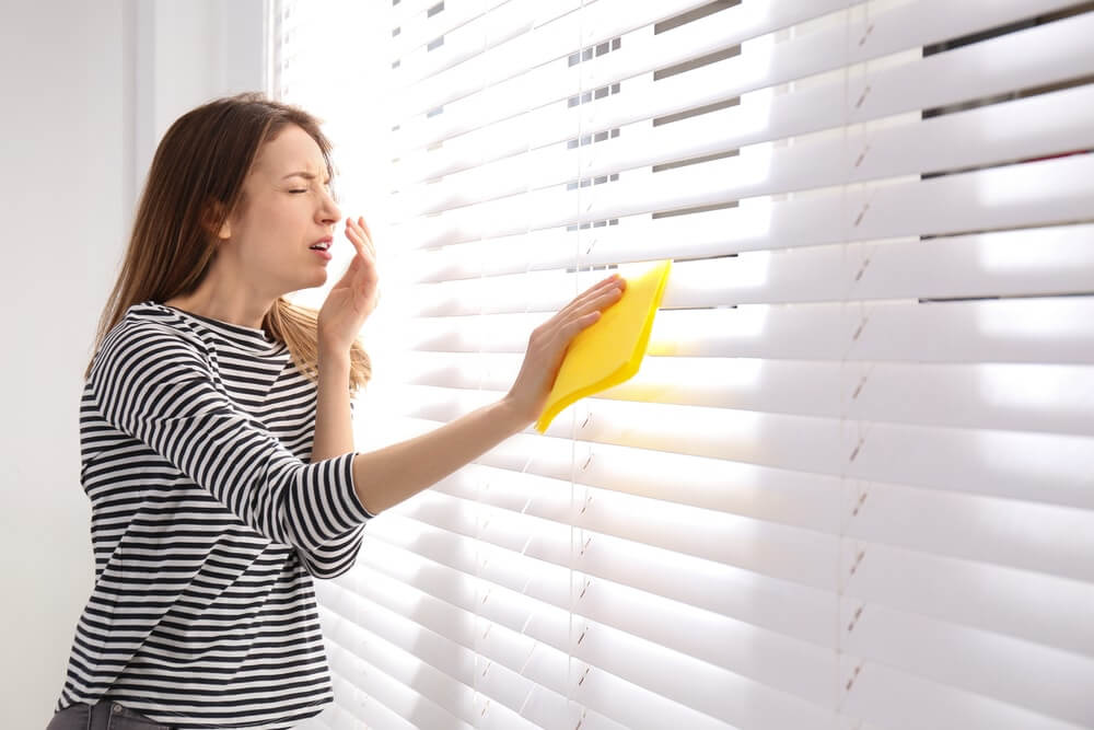 A woman in a striped shirt sneezes while wiping white blinds with a yellow cloth. The scene highlights the discomfort of dust, a common issue during home renovations like floor sanding and refinishing, which can affect air quality if not properly managed.
