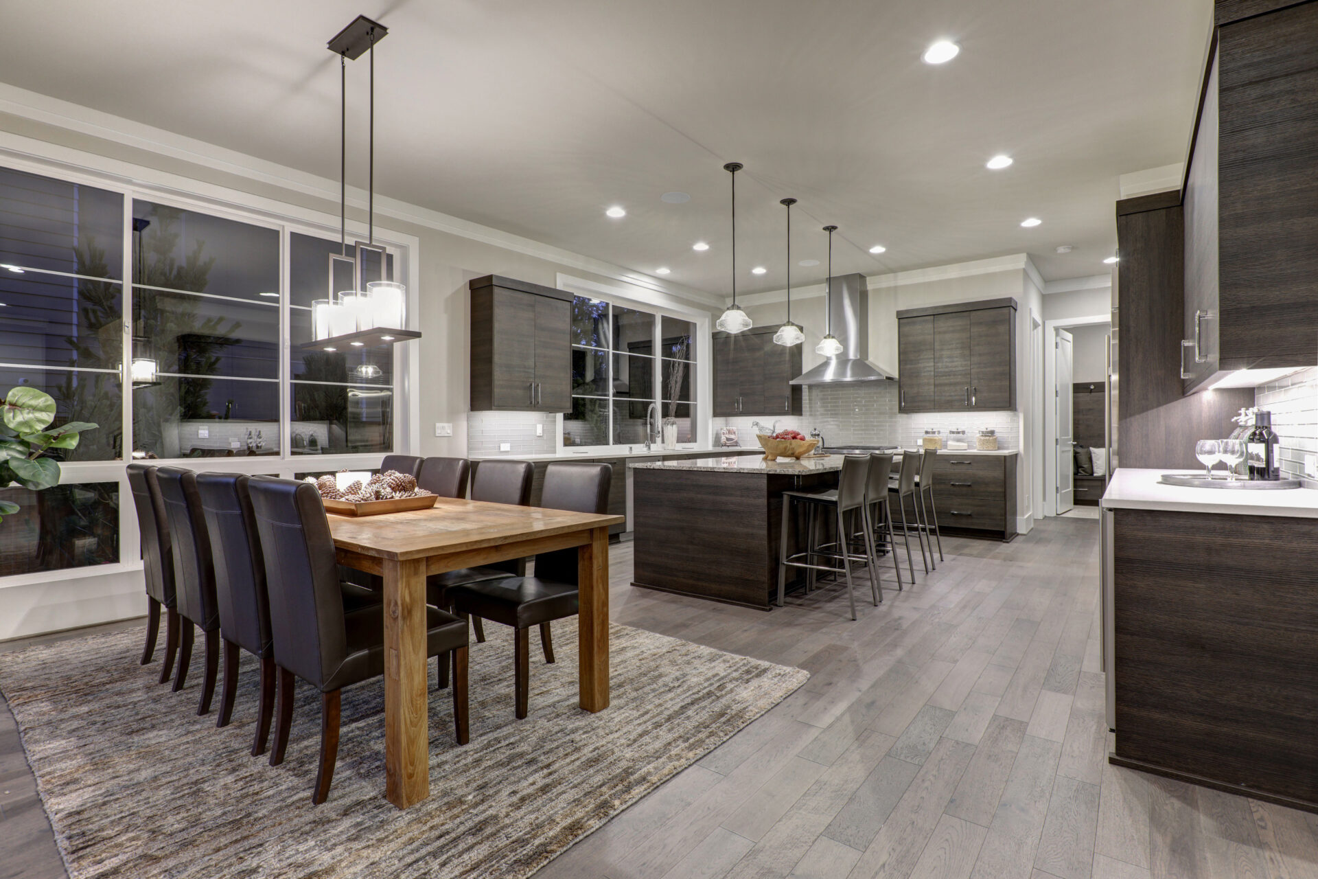  Modern open-plan kitchen-diner featuring grey hardwood flooring finished with Colourful Hardwax Oils, oak table and dark cabinetry under warm pendant lights.
