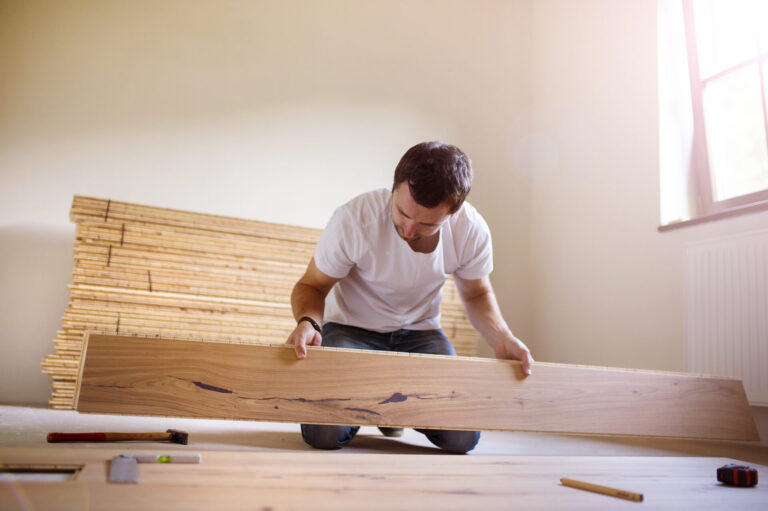 Male installer kneeling while fitting an oak floorboard, illustrating floor installation for noise reduction in a bright room.