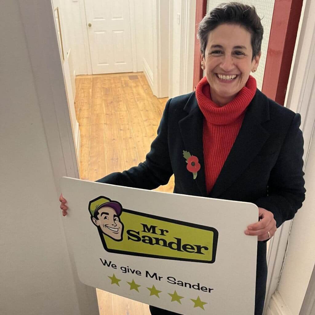 A smiling customer holding a "Mr Sander" sign with a 5-star rating, standing in a hallway with beautifully restored wooden floors in the background. The image highlights satisfaction with professional floor sanding services.