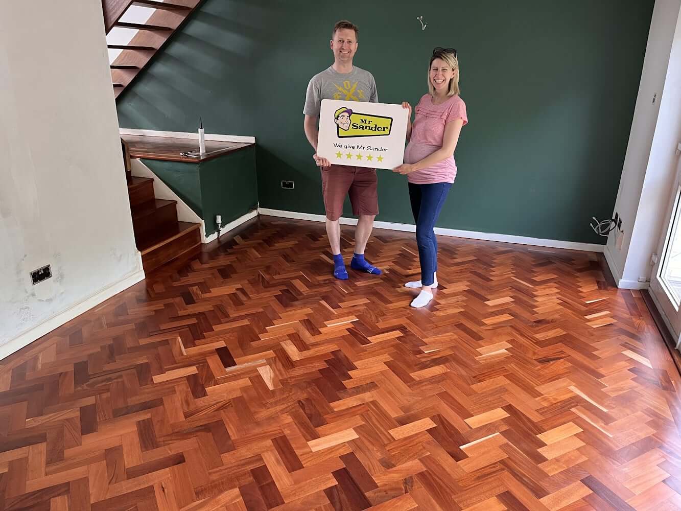 A happy couple standing on their newly sanded and refinished herringbone mahogany flooring done by Mr Sander® in UK. They are holding a Mr Sander® sign, showcasing their satisfaction with the professional floor restoration services. The room features a green accent wall, a staircase, and ample natural light highlighting the rich tones and intricate pattern of the restored mahogany floor.