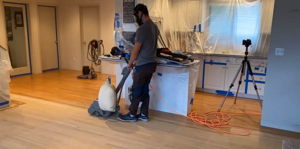 A professional floor refinisher uses a sanding machine to refinish a light oak hardwood floor. The room is under renovation, with plastic sheets covering the kitchen cabinets and equipment to protect against dust. A tripod with a camera is set up in the background, documenting the process. The refinishing project takes place in an open-plan space, with the hardwood floor being prepped for a smooth and polished finish.