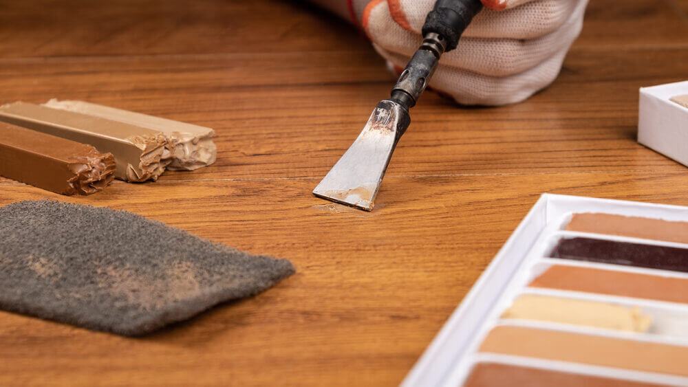 A gloved hand uses a scraping tool to apply a colour-matched wax fill on a wooden floor during a Wood Floor Scratch Repair process.