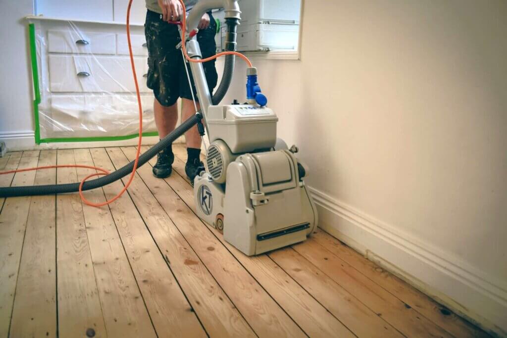 Man from Mr Sander® sanding a pine floor in a living room in Colnbrook, UK, using a Bona Belt Lite (AMK175000) belt sander. Technical specs: 2.2 kW motor power, 230 V, 50 Hz, 200x750 mm sanding belt size, 200 mm drum width, 2400 rpm drum revolutions, 60 kg total weight.