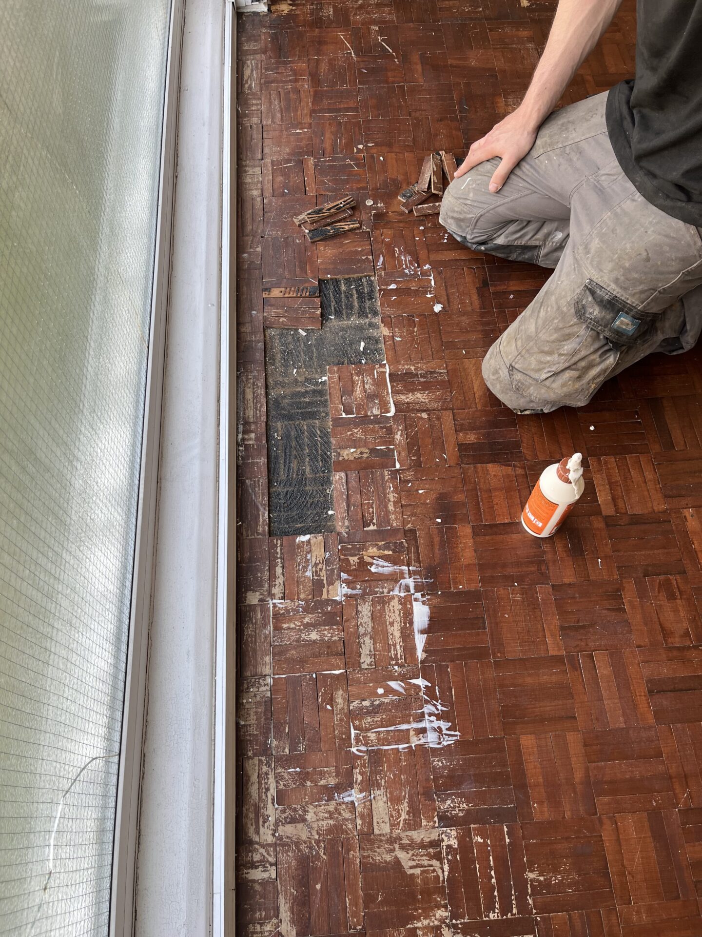 A tradesperson kneeling on a dark mosaic parquet floor with a worn finish, carefully replacing damaged blocks near a window using wood adhesive.
