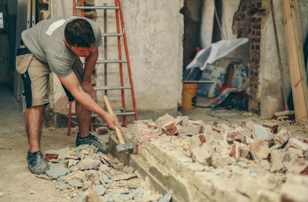 A construction worker using a sledgehammer to break down rubble in a partially demolished room. The background shows a ladder and various construction materials.