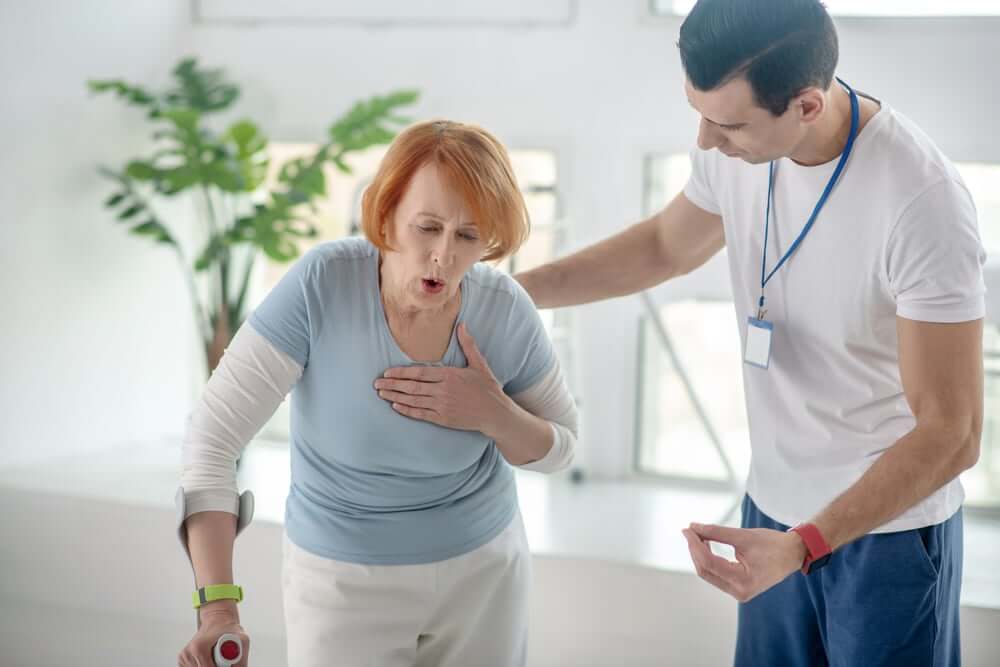 An older woman coughing and holding her chest, assisted by a healthcare professional, illustrating respiratory distress that may be linked to inhaling dust particles.
