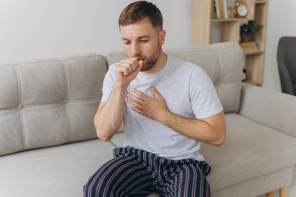 A young man coughing into his fist while placing a hand on his chest, suggesting respiratory irritation possibly linked to dust inhalation.