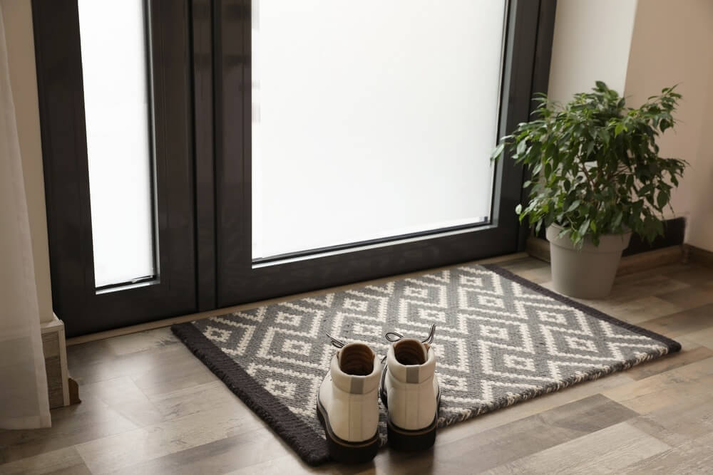 Entrance area with a doormat and a pair of white shoes placed on it, adjacent to a potted plant. The newly refinished wooden floor by Mr Sander® shines, showcasing its smooth and polished finish.